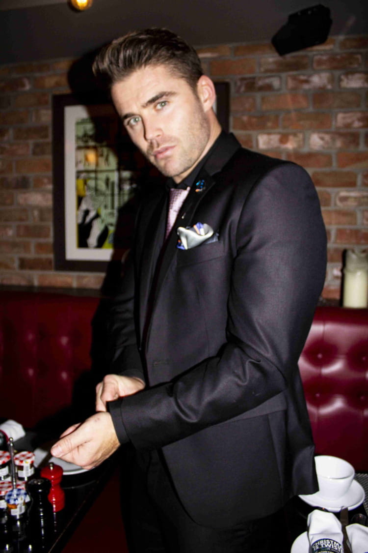 Man in a black suit with a red brick wall and framed picture in the background