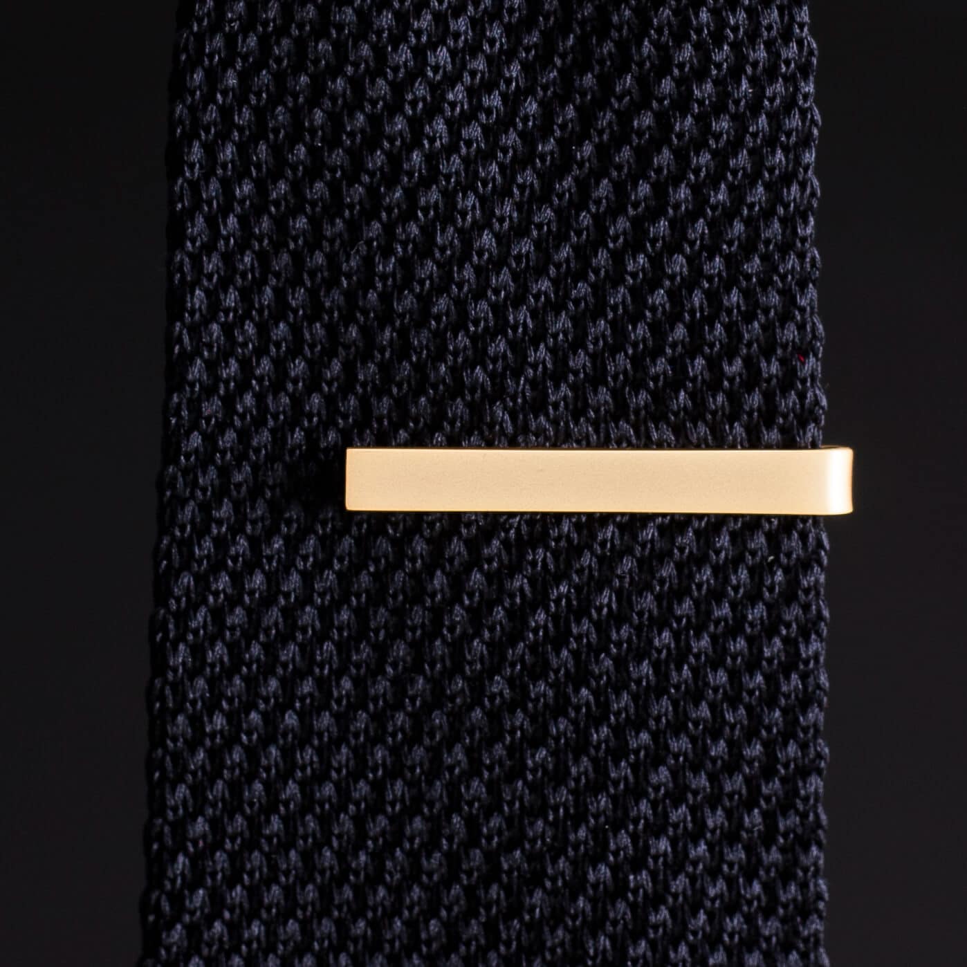 Navy patterned tie with a gold tie clip on a black background