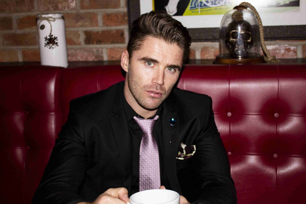 Man in a suit sitting in a red booth holding a cup, with a brick wall and framed picture in the background.