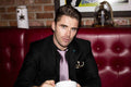Man in a suit sitting in a red booth holding a cup, with a brick wall and framed picture in the background.