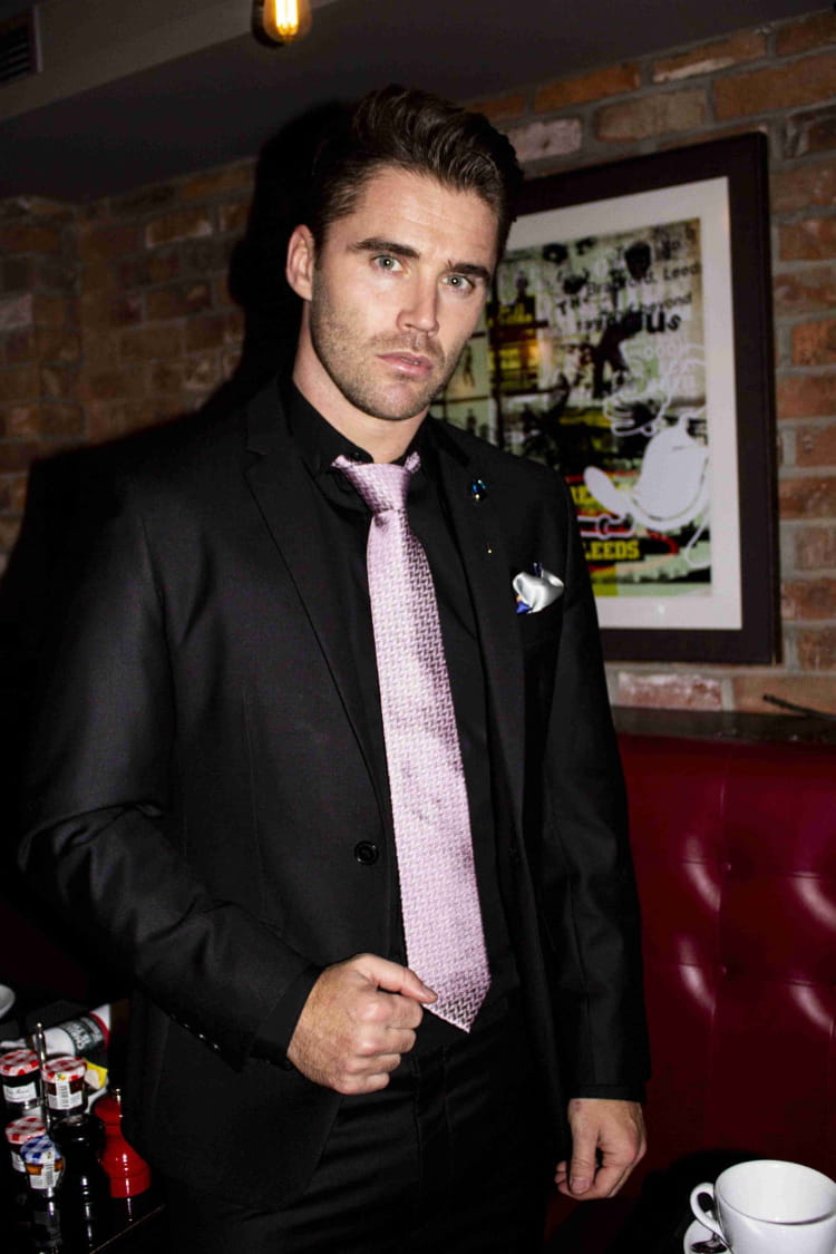 Man in a black suit with a pink tie standing in a room with a brick wall and framed picture.
