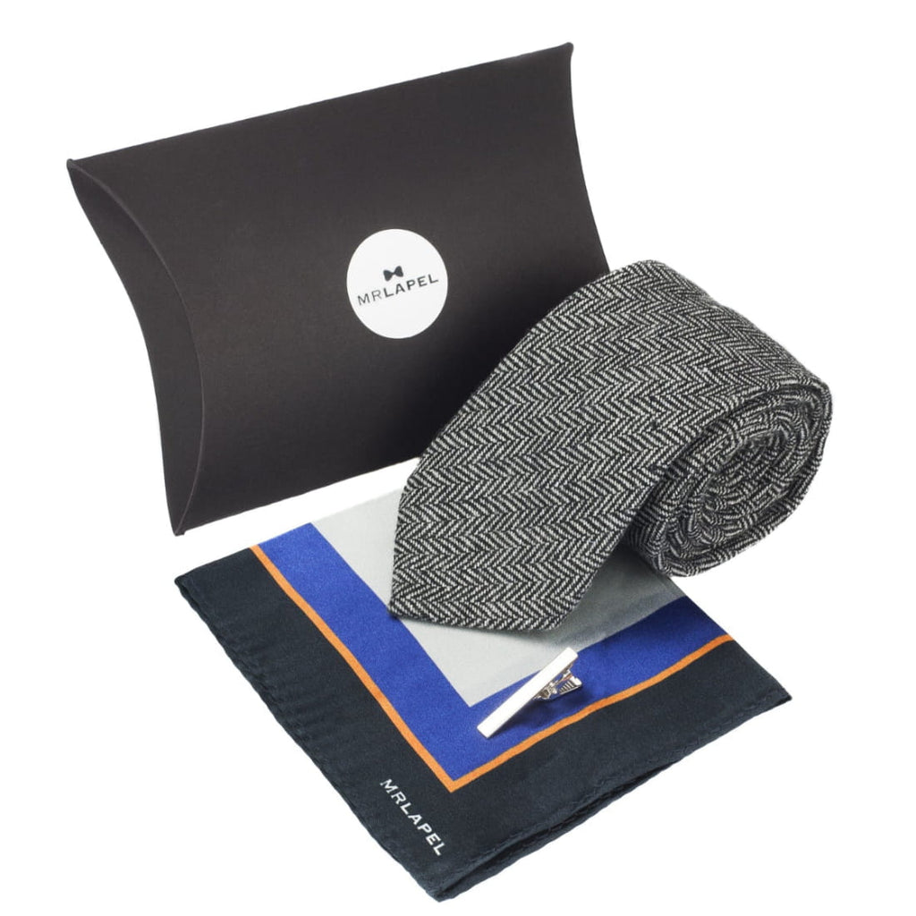 Gift set featuring grey herringbone tie, printed silk pocket square with mix of grey, black, orange and blue. and a silver tie clip, with large black presentation box behind it