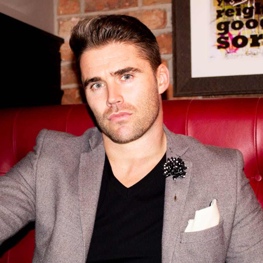 Man in a gray blazer with a black shirt and floral brooch sitting on a red couch.