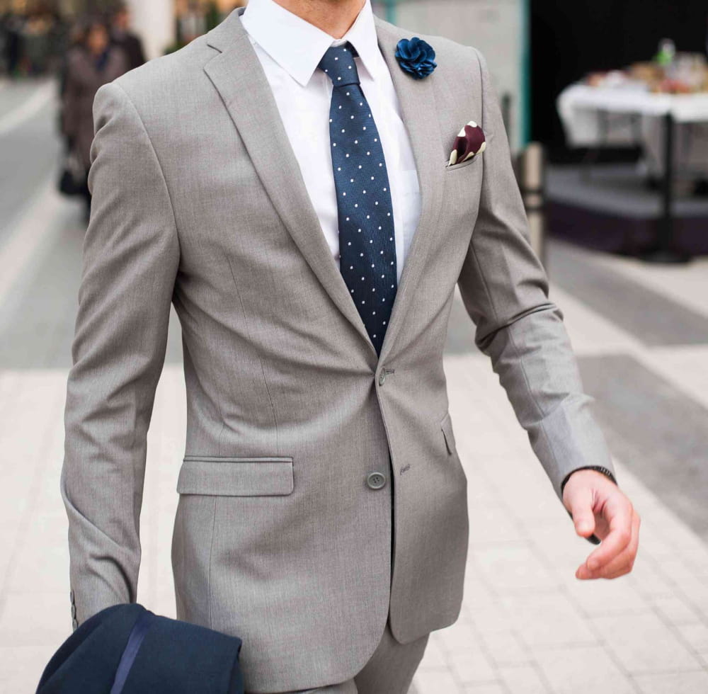 Man wearing a gray suit with a blue tie and pocket square on a city street.