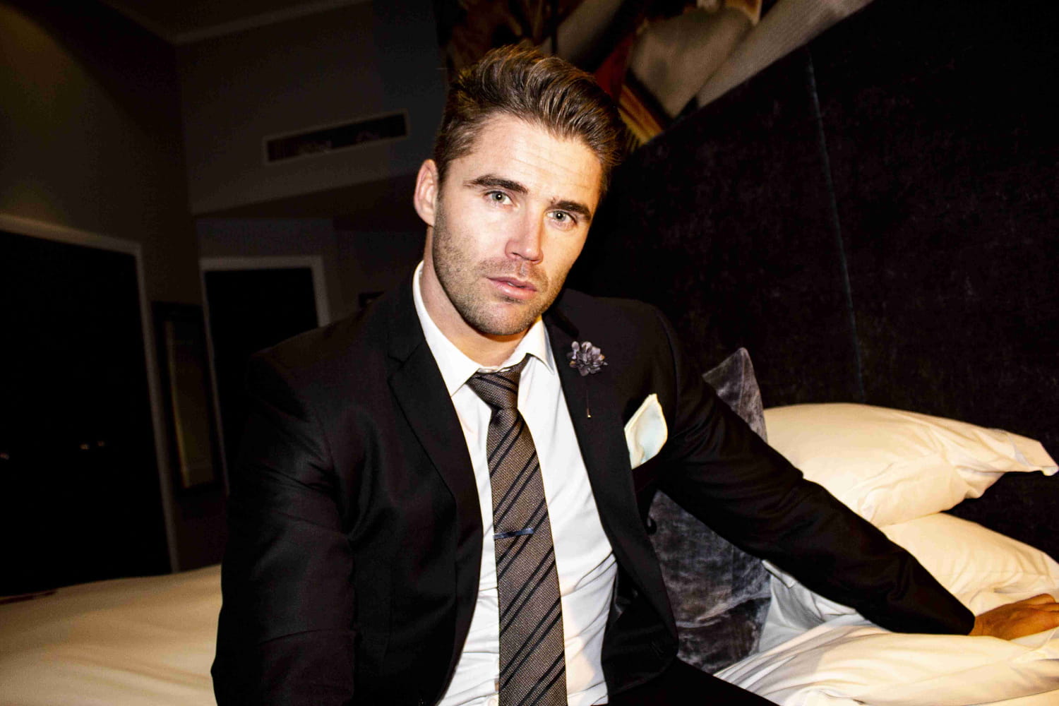 Man in a suit sitting on a bed in a dimly lit room